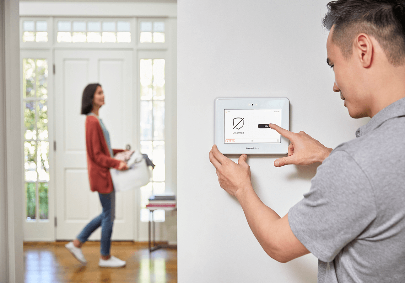 Person adjusts settings on a wall-mounted tablet while another person, carrying a bag, walks in the background of a bright room with a front door.